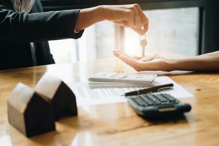 Person handing over house keys to another person during a real estate transaction, with cash, documents, calculator, and small wooden house models on a wooden table.